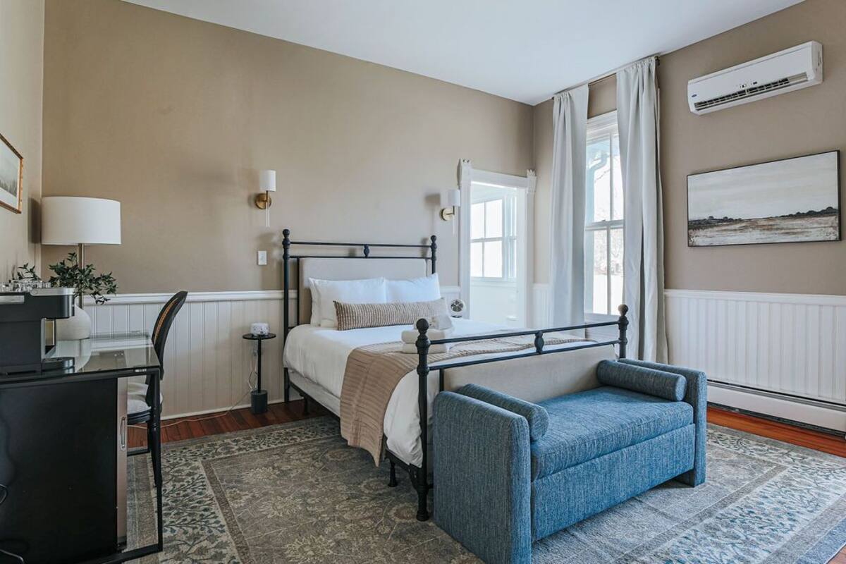 A bedroom with a bed, bedside table, lamp, chair, and a large window with curtains.