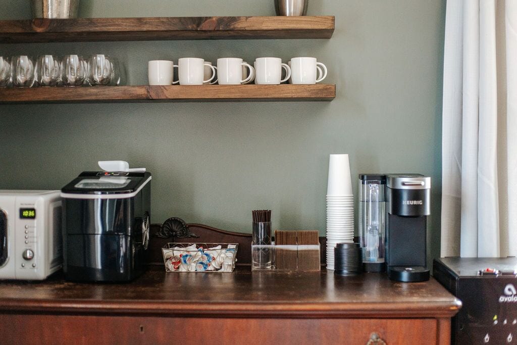 A kitchenette with a coffee maker, a toaster, and a microwave. There are also stacked white cups and a box of tea bags.