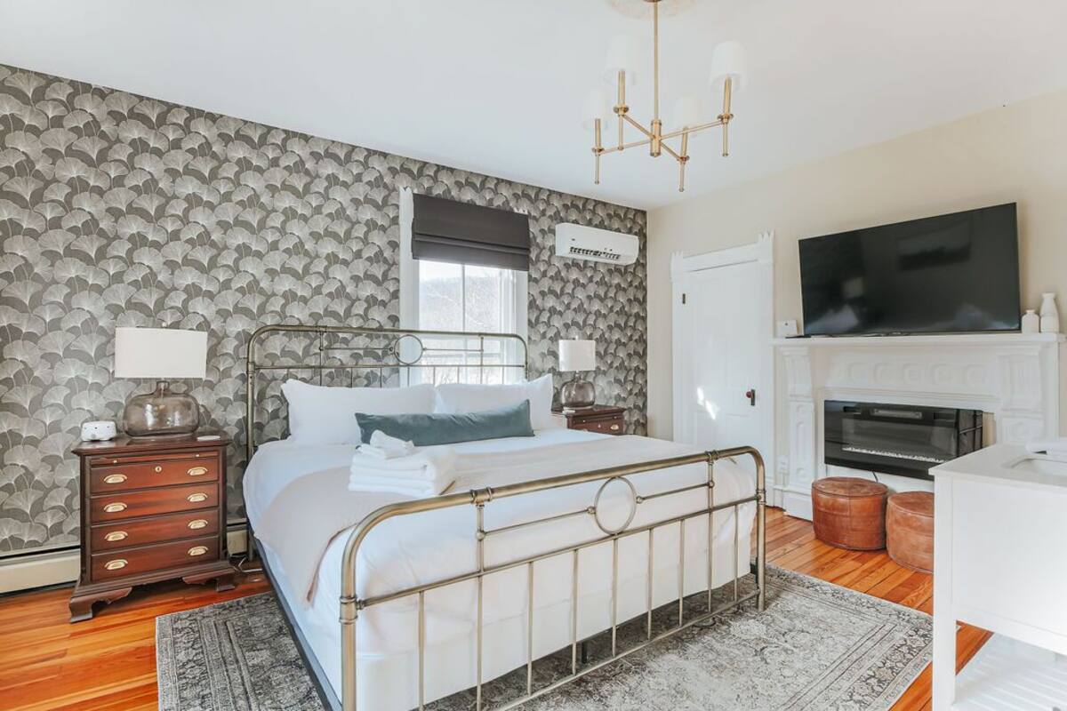 A bedroom with a bed, nightstands, a TV, a fireplace, and patterned wallpaper.