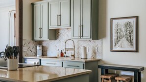 A kitchen with green cabinets, a herringbone-patterned backsplash, a central island, and a wooden stool bench.