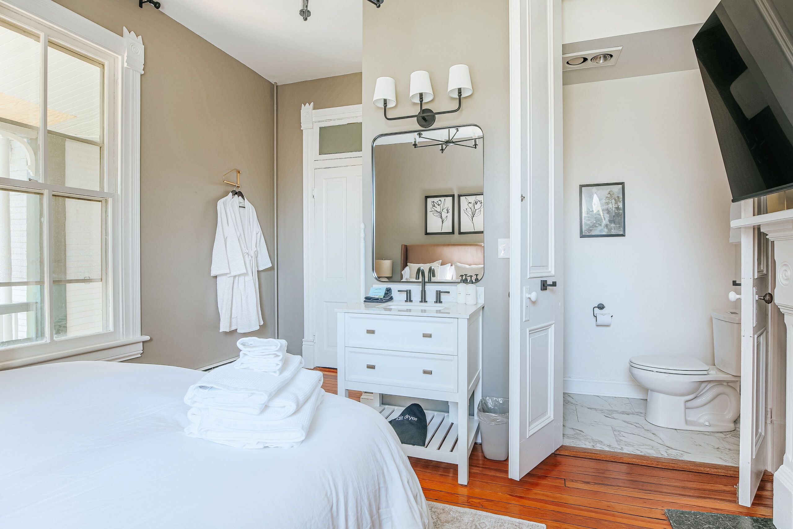 Une chambre à coucher comprenant un lit, une commode blanche, une coiffeuse avec un miroir, et une salle de bain avec des toilettes et un lavabo.