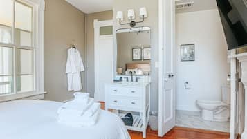 Une chambre à coucher comprenant un lit, une commode blanche, une coiffeuse avec un miroir, et une salle de bain avec des toilettes et un lavabo.