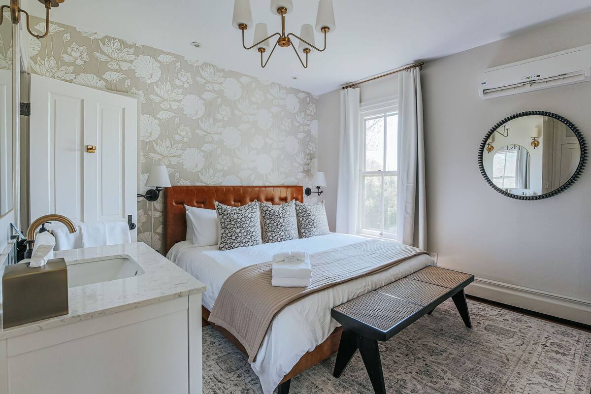 A bedroom with a large bed, a bedside table, a mirror, a window with curtains, a chandelier, and an air conditioning unit.