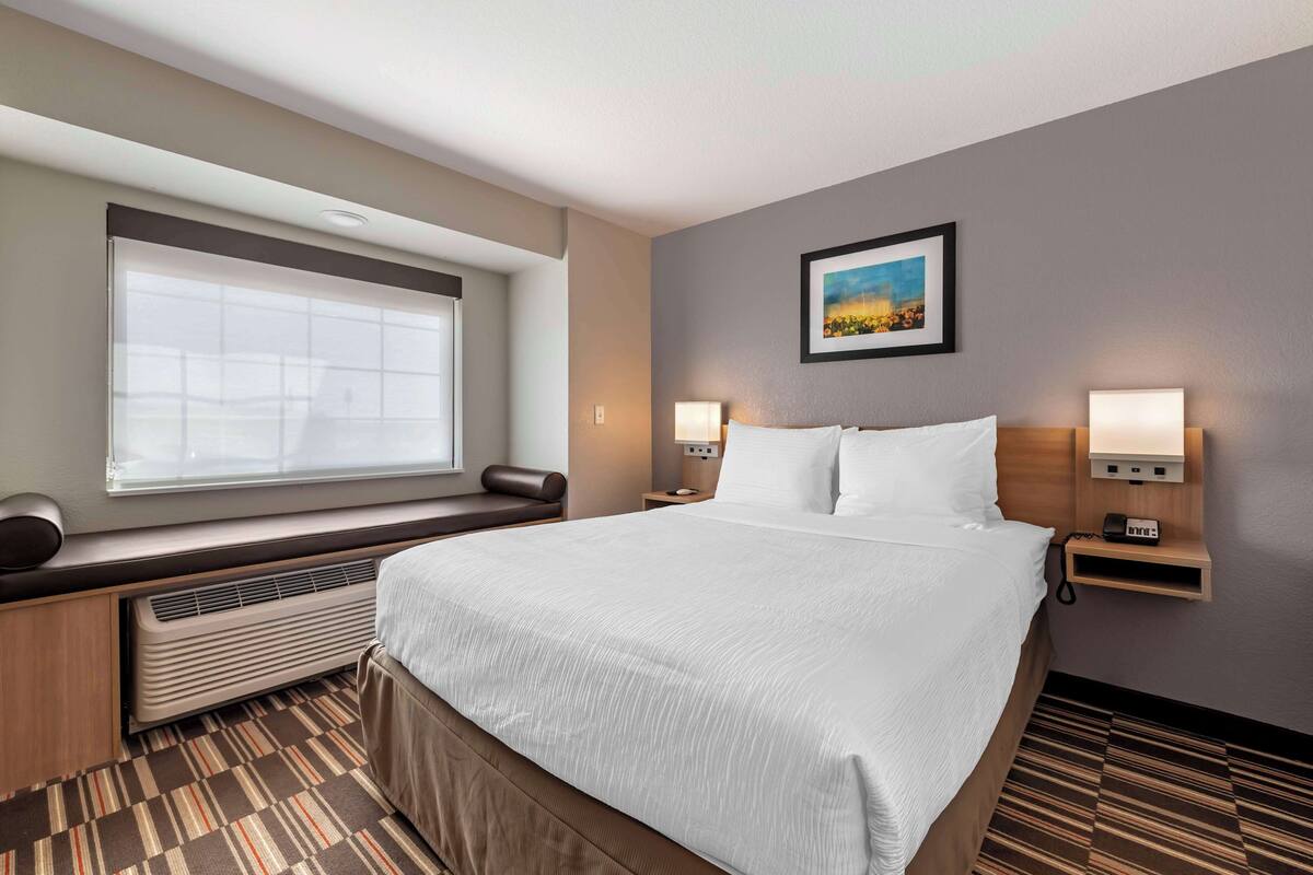 A hotel room with a large bed, a window with blinds, a wall-mounted picture, two bedside lamps, and a built-in heater.