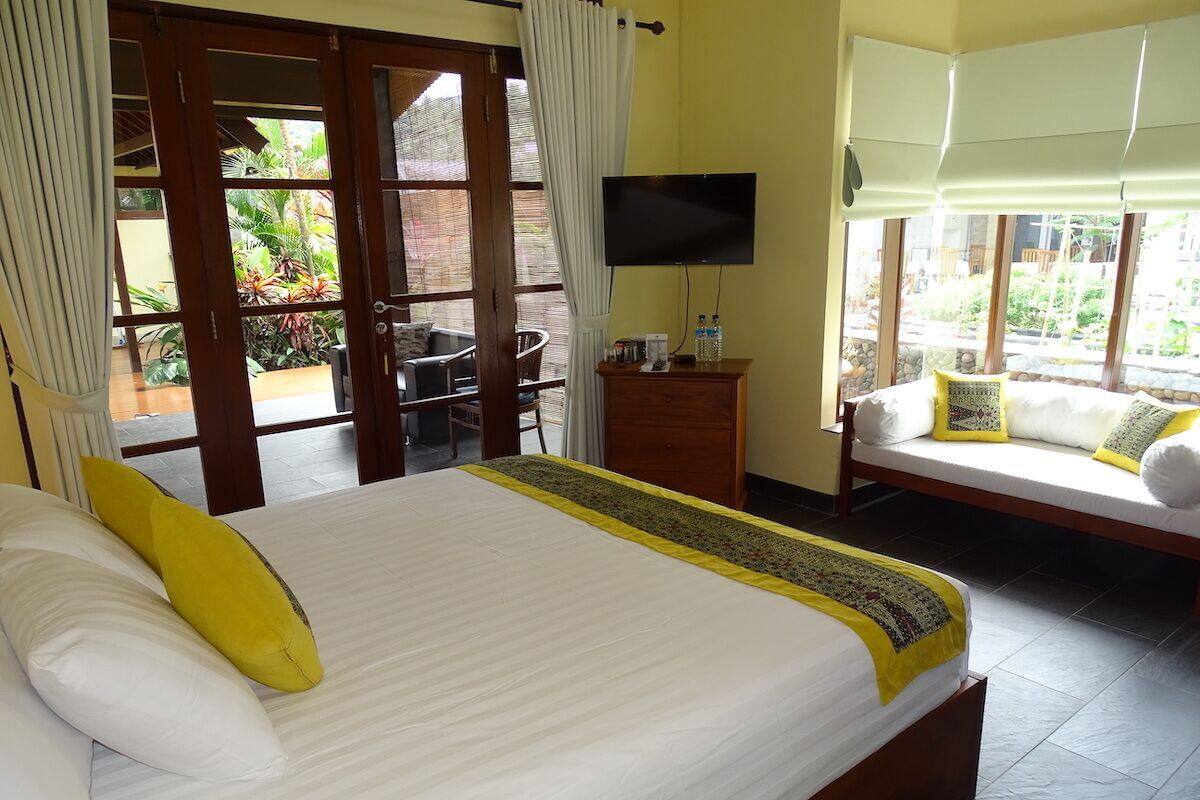 A hotel room with a large bed, a TV mounted on the wall, a sofa with cushions, and a view of the garden through the window.
