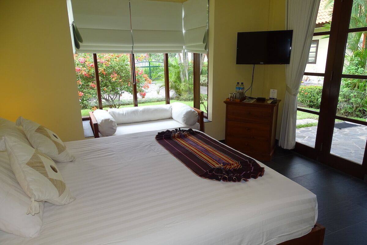 A hotel room with a large bed, a television, a wooden dresser, and a view of a garden through the window.