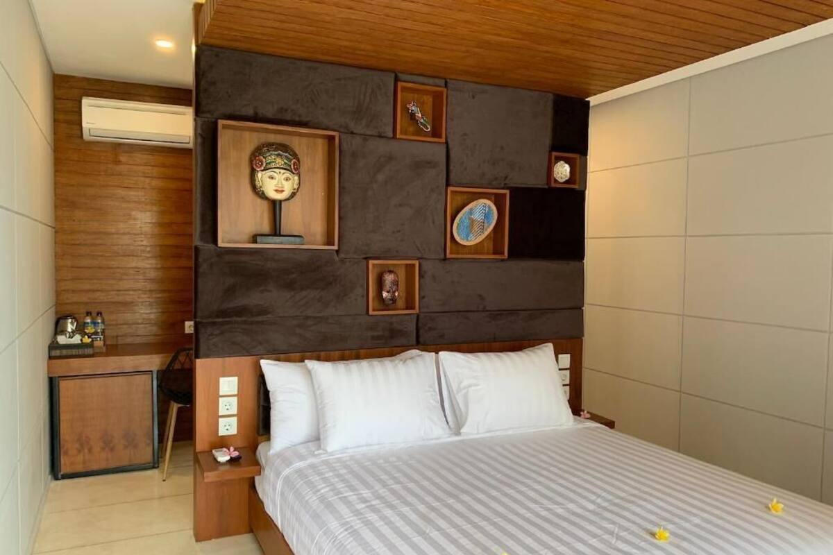 A hotel room with a wooden headboard, a bed with white bedding, a small bedside table, and a wall-mounted shelf with decorative items.