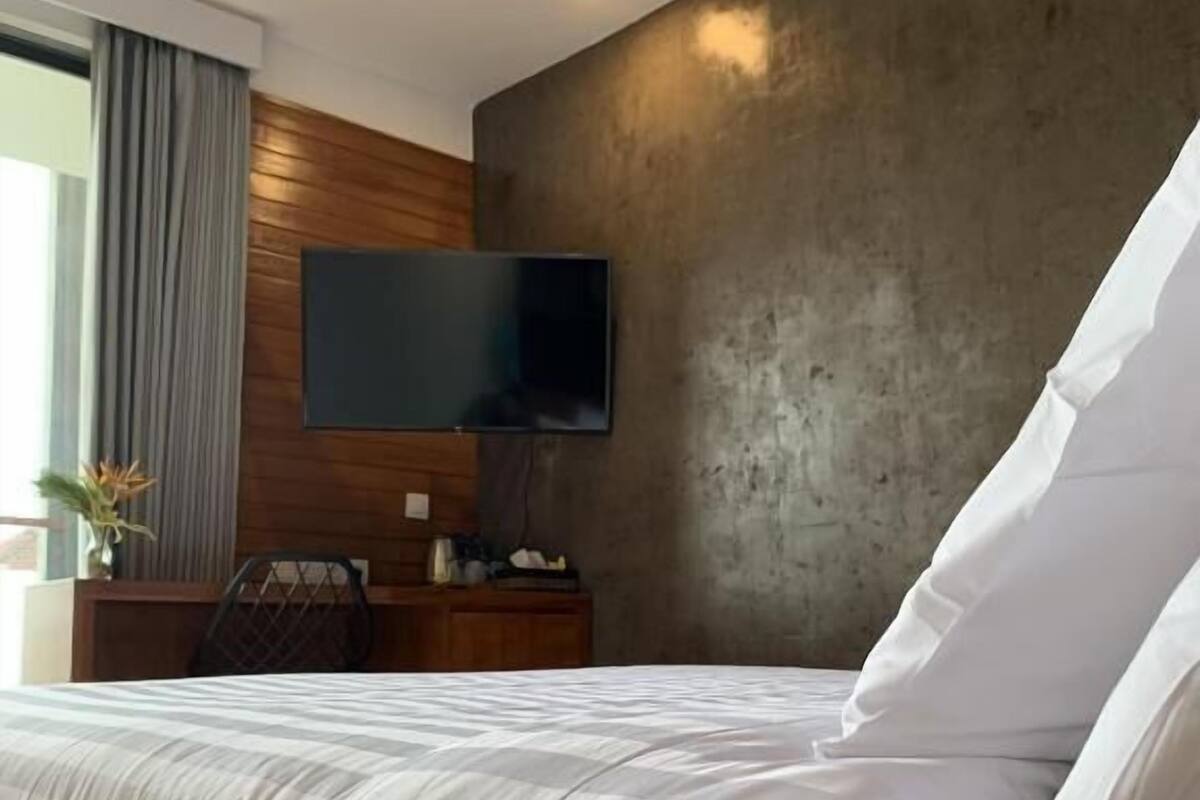 A modern hotel room with a large bed, a flat-screen TV mounted on a concrete wall, and a wooden ceiling.