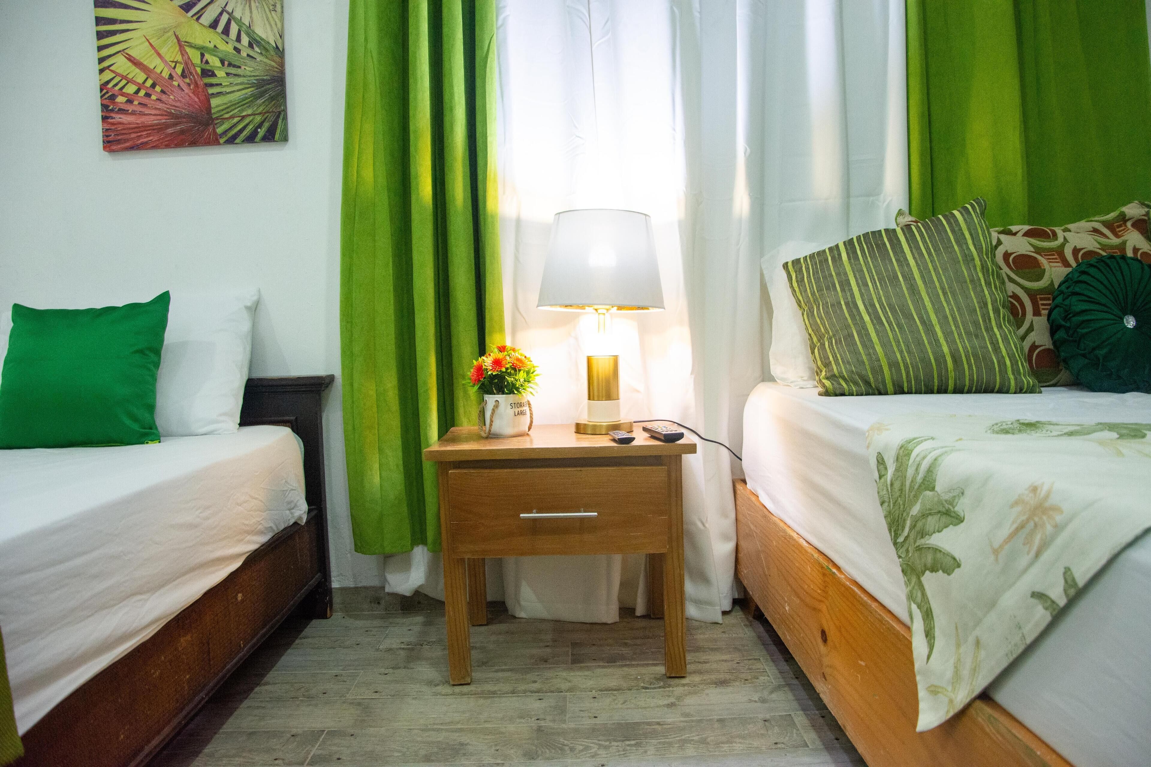 Une chambre d’hôtel avec deux lits simples, une table de chevet avec une lampe, une petite table avec un vase de fleurs et des rideaux verts.