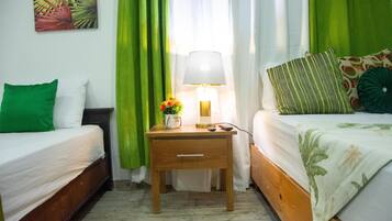 Une chambre d’hôtel avec deux lits simples, une table de chevet avec une lampe, une petite table avec un vase de fleurs et des rideaux verts.