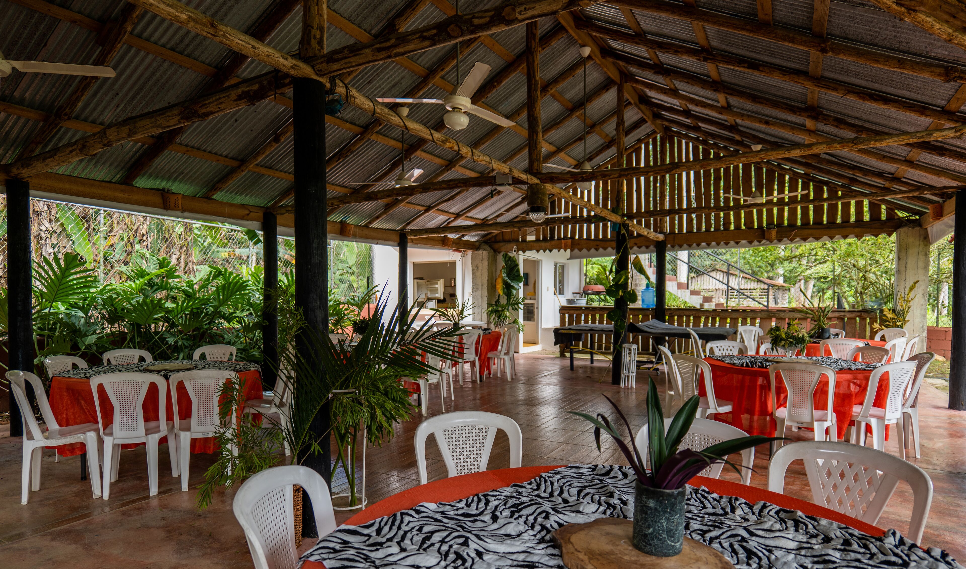 A covered outdoor dining area with tables and chairs, surrounded by lush greenery.
