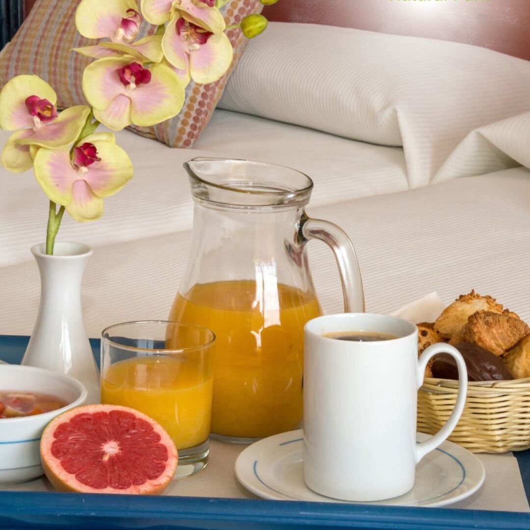A breakfast tray with orange juice, coffee, grapefruit, and pastries on a bed with pillows and an orchid vase.
