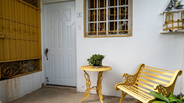 A white door with a number 4 sign, a barred window, a small round table with a plant, and a yellow bench. - Hotel Santo Cerro Natural Park  (La Vega)