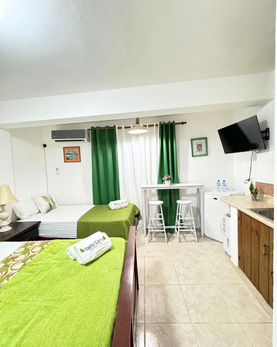 Een compacte hotelkamer met een bed, een kleine kitchenette, een bureau met twee krukken en een aan de muur bevestigde televisie.