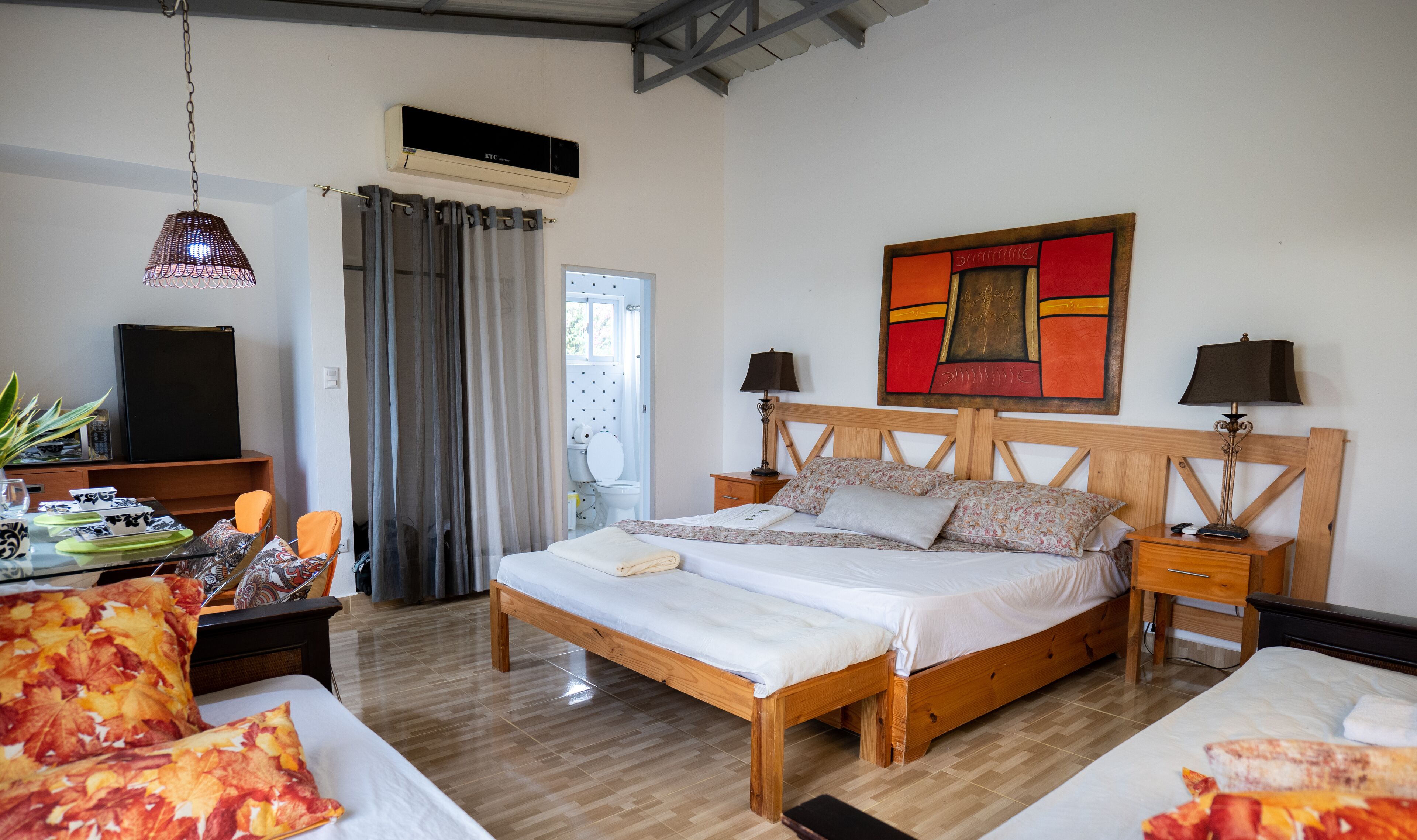 Un dormitorio con cama de madera, dos mesitas de noche con lámparas, un escritorio con silla, una lámpara colgante, un cuadro en la pared y una ventana con cortinas.