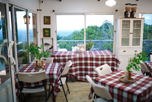 A dining area with tables covered in plaid tablecloths, white chairs, and a view of greenery through a glass door.
