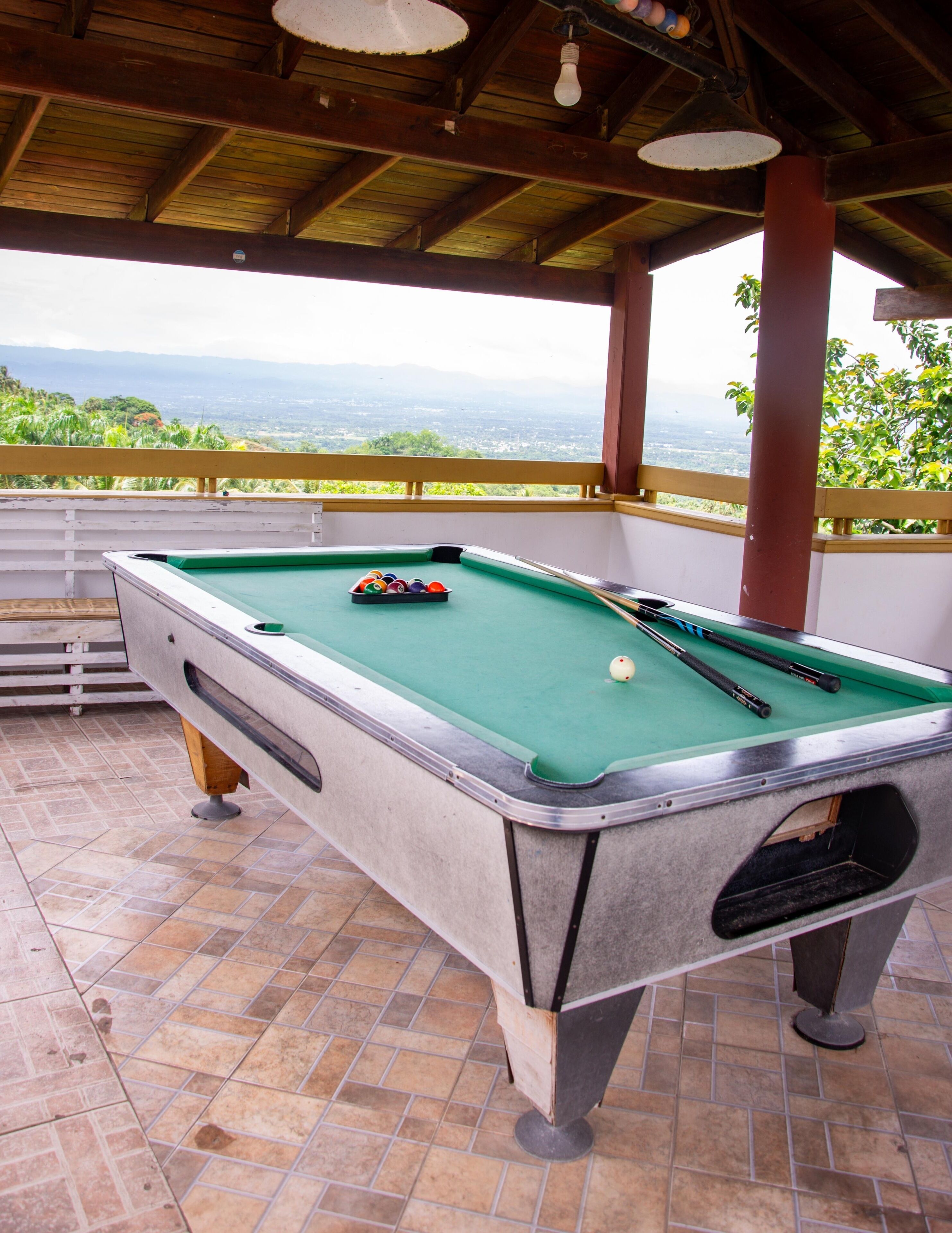 A pool table with balls and cues, set on a tiled floor under a wooden ceiling with a view of greenery and mountains in the background.