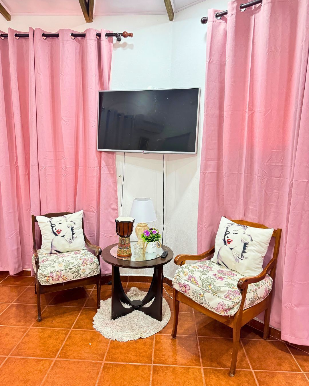 Una habitación con dos sillas de madera tapizadas con motivos florales, una mesa redonda y una alfombra blanca, un televisor montado en la pared y cortinas rosadas.
