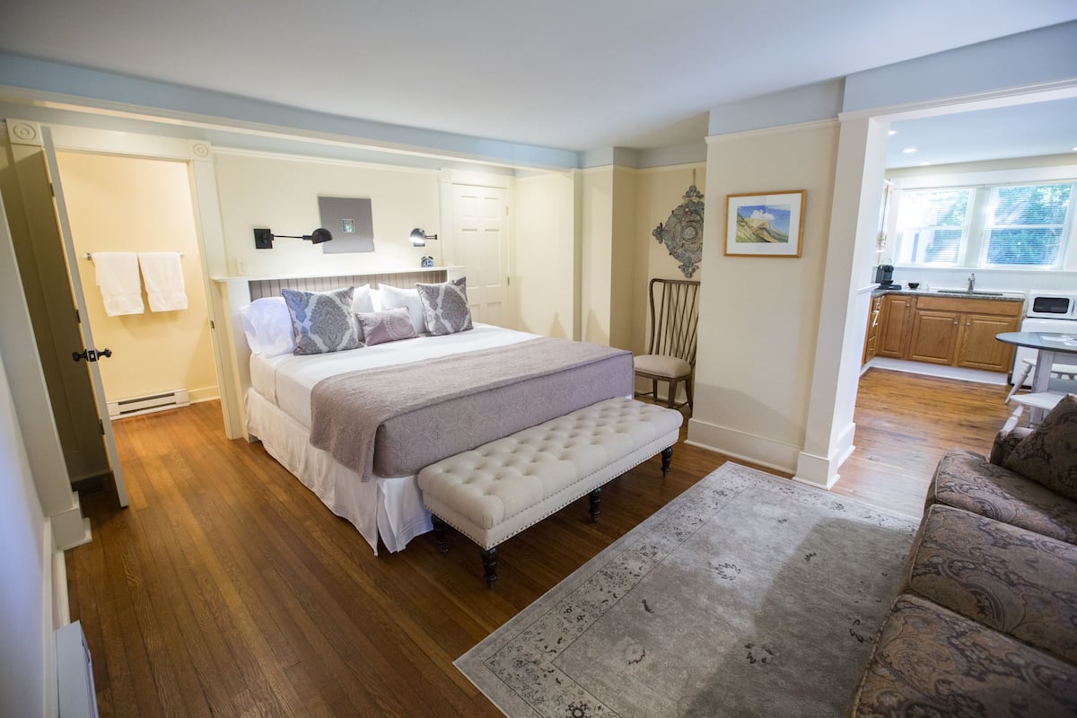 A bedroom with a bed, bench, and a view of the kitchen.