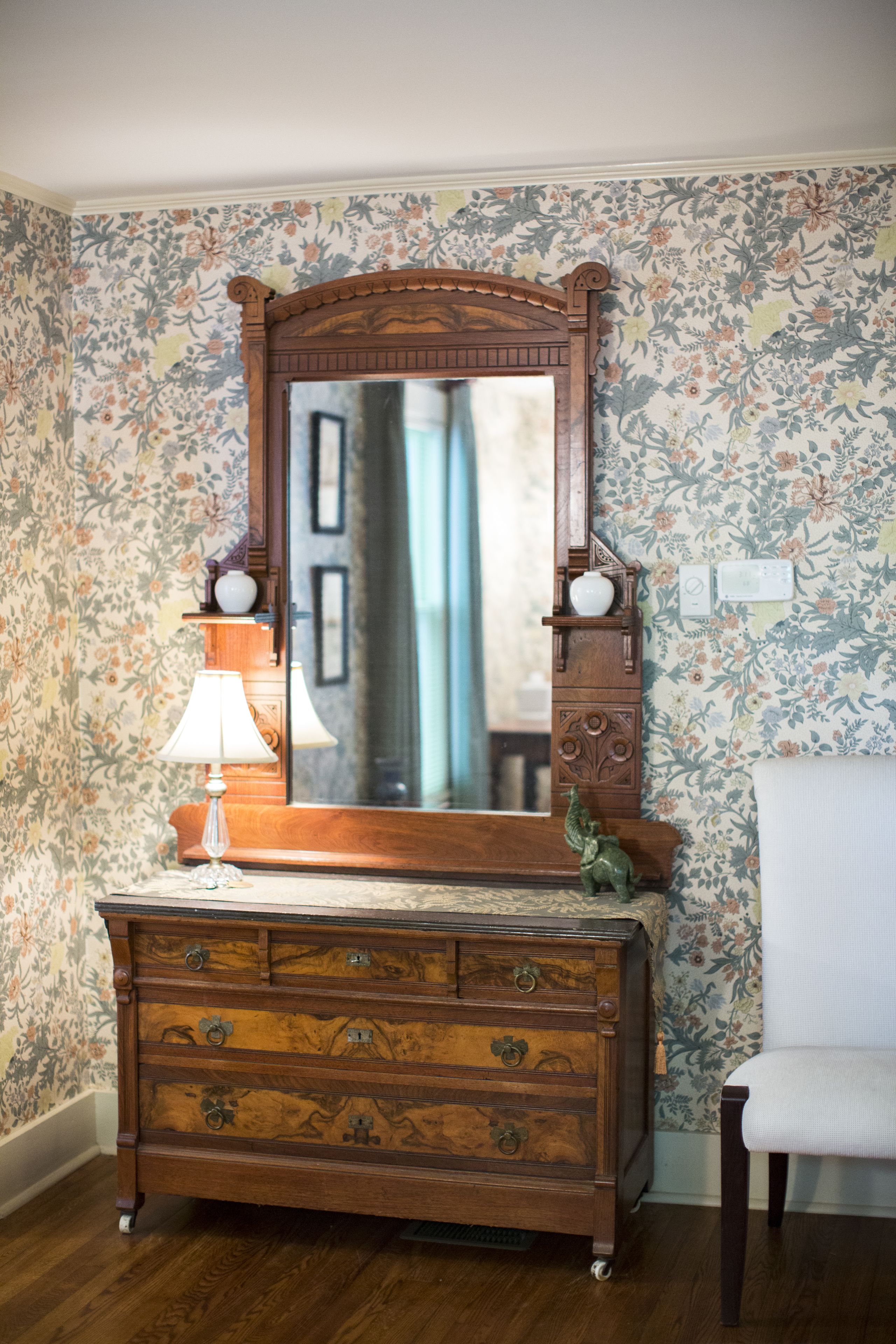 A wooden dresser with a mirror above it, a lamp on the dresser, and a chair to the right.