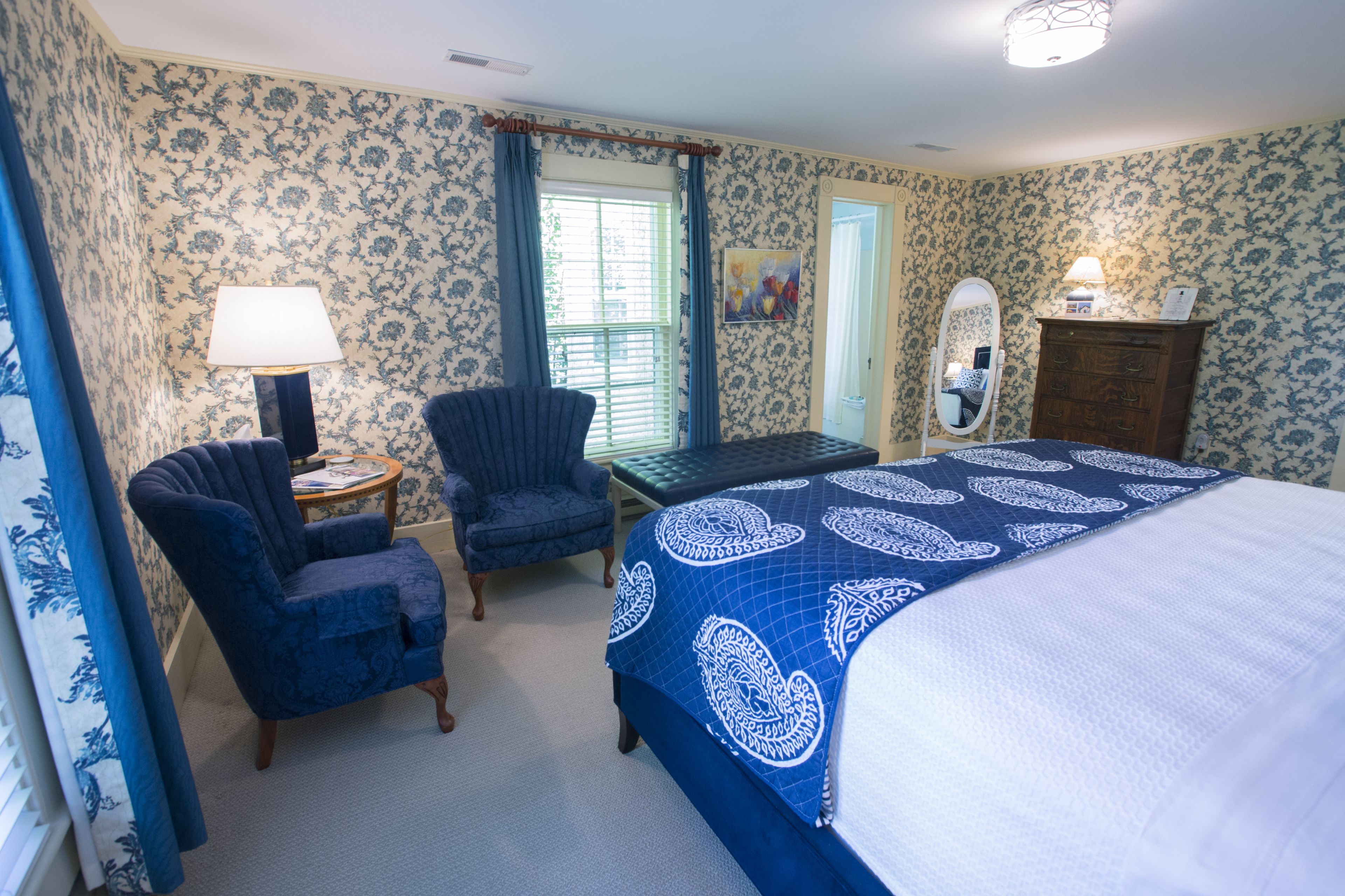A bedroom with a bed, two armchairs, a table, a dresser, a mirror, and a window with curtains.