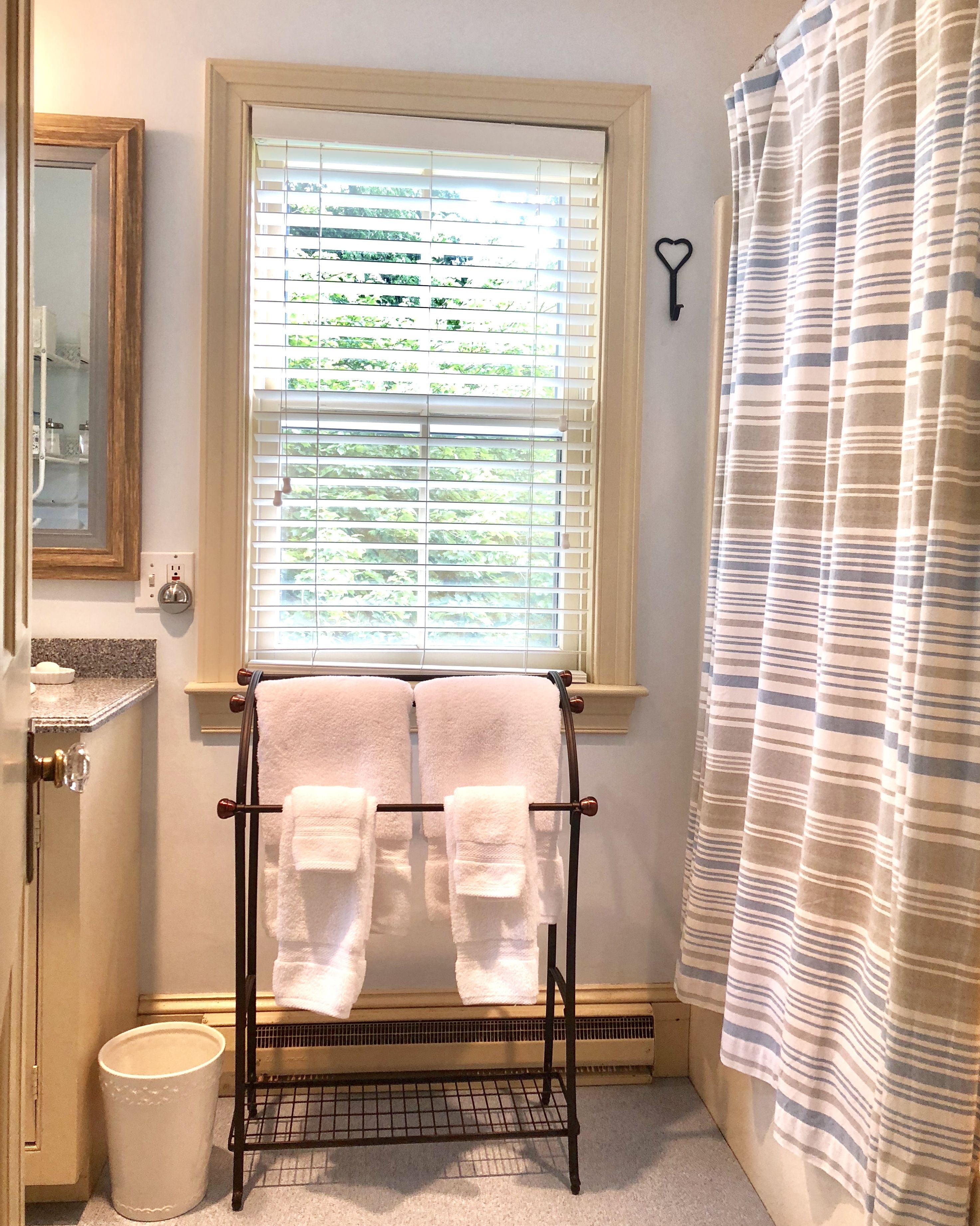 A bathroom with a window, a towel rack with two towels, a shower curtain, a mirror, and a sink with a soap dispenser.