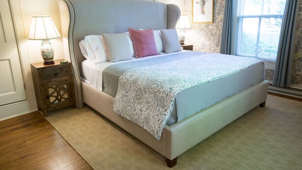 A bedroom with a large bed, a nightstand with a lamp, a wooden side table, and a window with curtains.