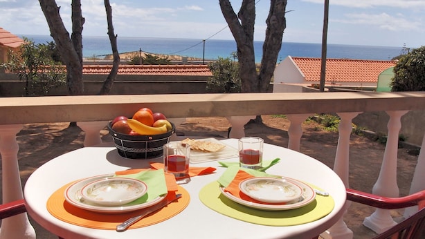 Una mesa dispuesta con platos, tazones, un frutero y vasos en un balcón con vistas al mar.