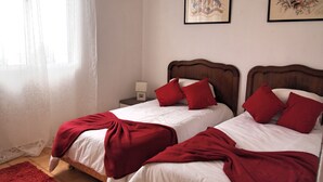 Habitación de hotel con dos camas, cada una con cabecero de madera, ropa de cama blanca y almohadas rojas. Hay una mesita de noche con lámpara entre las camas y una ventana con cortinas de gasa.
