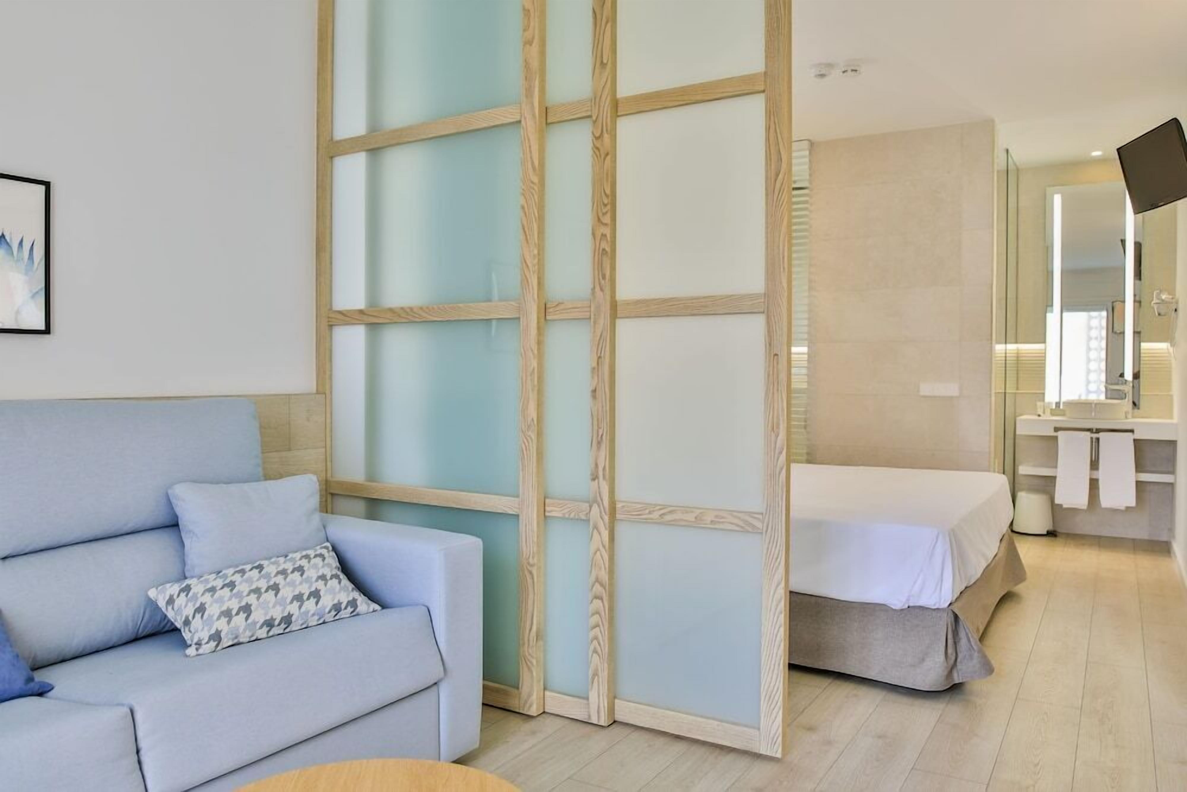 A modern hotel room with a light blue sofa, a bed with a wooden headboard, a large mirror, and a bathroom with a sink and mirror.