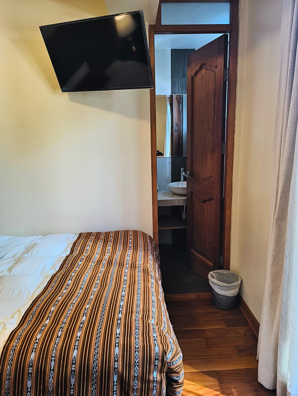 A bedroom with a bed, a television mounted on the wall, a wooden door leading to a bathroom, and a small trash bin.