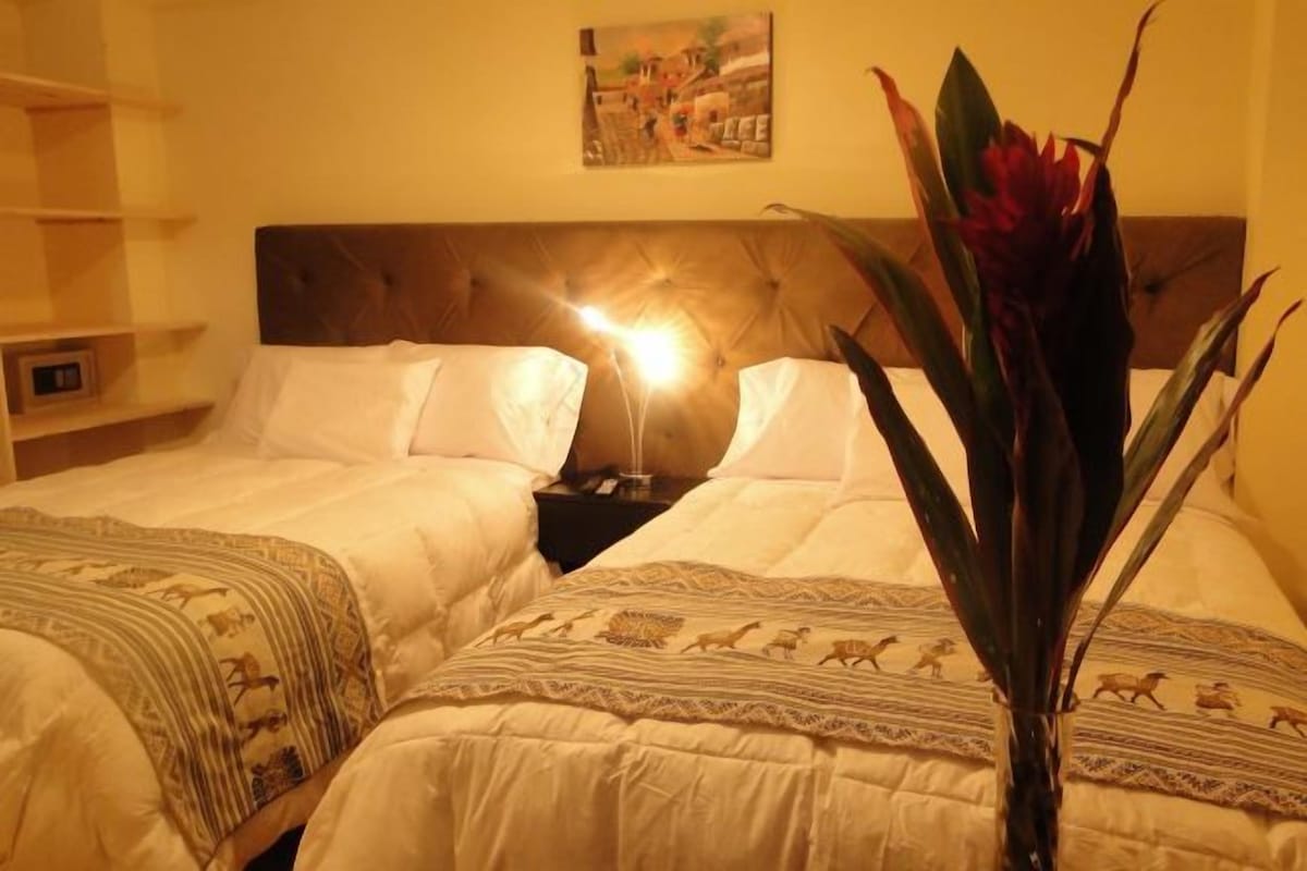 A hotel room with two beds, a nightstand, a lamp, and a large plant.