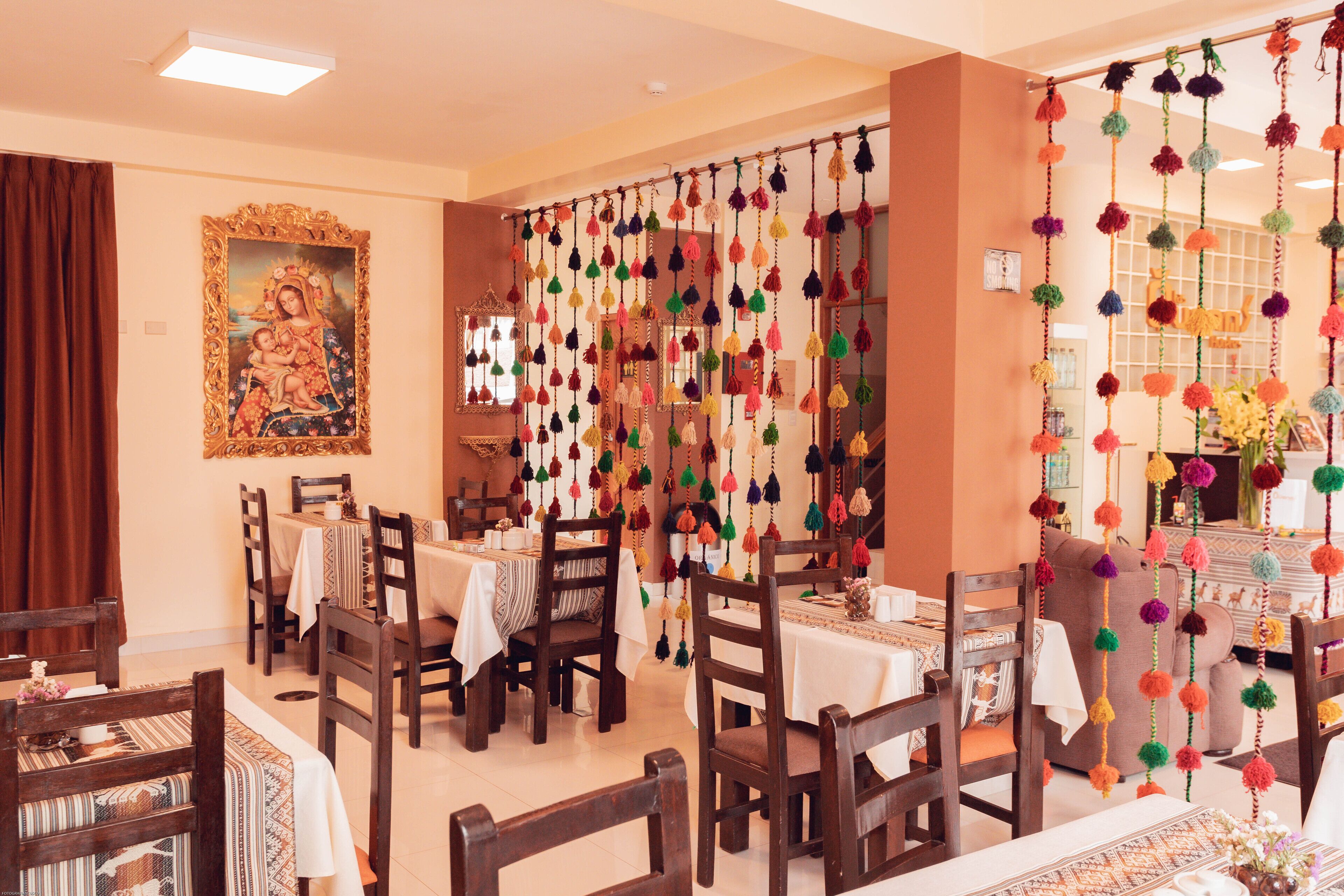 A dining area with wooden chairs and tables, a large framed painting on the wall, and colorful hanging decorations.