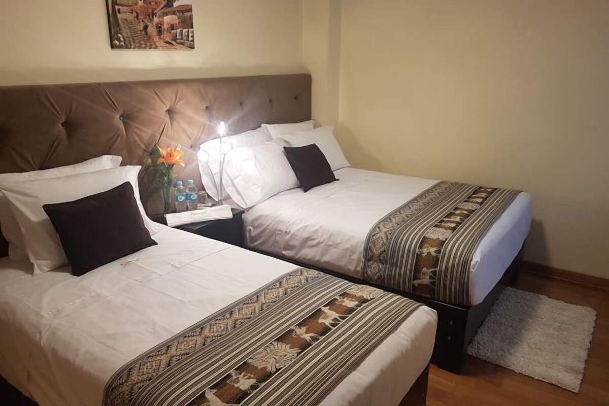 Two twin beds with white linens and dark pillows, a nightstand with a lamp, and a decorative vase with flowers.