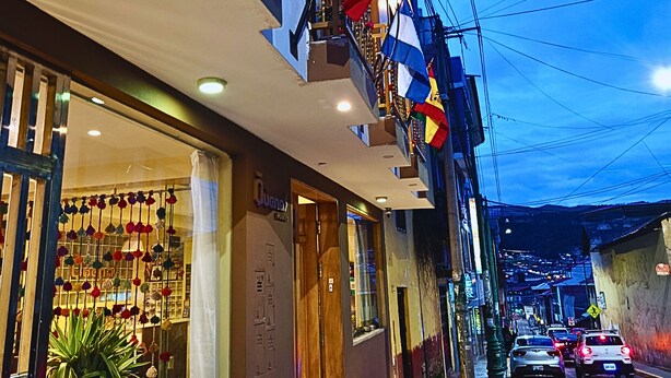 A street view at dusk with a building featuring balconies with flags, a storefront with a display of flowers, and parked cars along the road. - Hotel Las Quenas (Cusco)