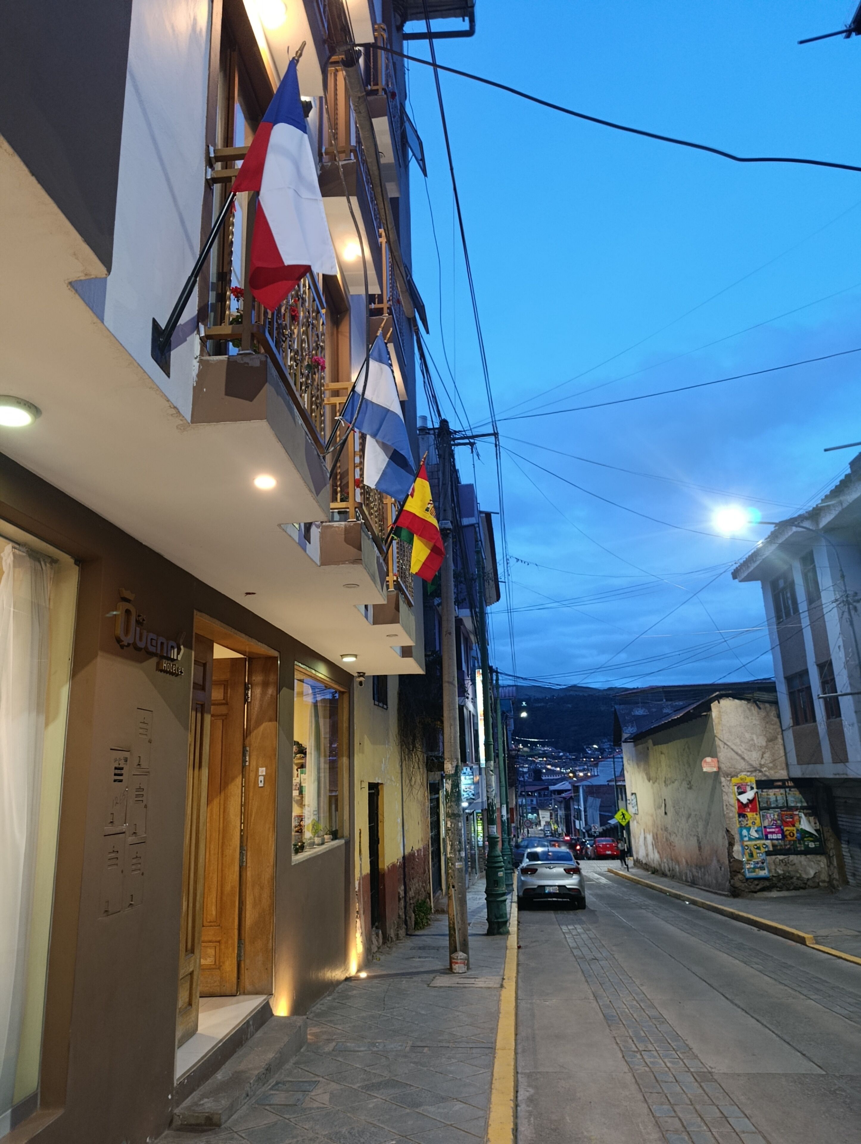 A street view with a hotel named 'Quenns' on the left, a balcony with flags, and a clear sky.