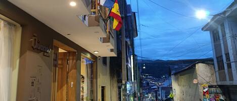 A street view with a hotel named 'Quenns' on the left, a balcony with flags, and a clear sky.