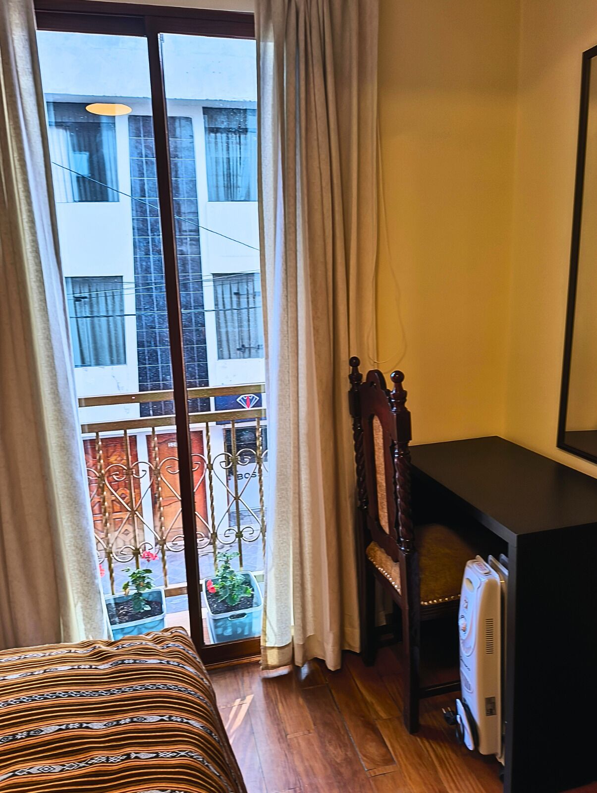 Habitación con cama, escritorio y radiador. Se ve una ventana con cortinas y vistas a edificios.