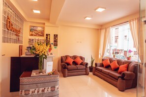 A modern hotel lobby with a reception desk, two sofas with orange cushions, a patterned ottoman, a vase with yellow flowers, and a large window with a city view. - Hotel Las Quenas (Cusco)