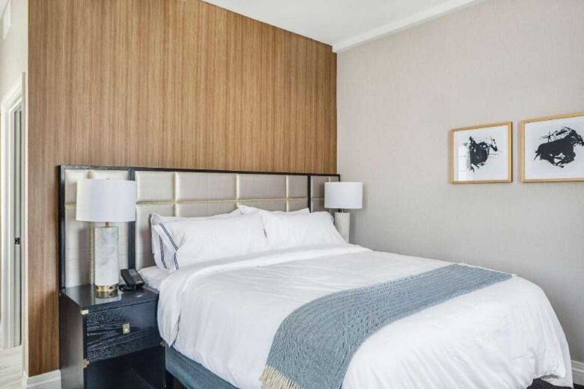 Habitación de hotel con cama, mesitas de noche, lámpara, una silla y dos cuadros en la pared.