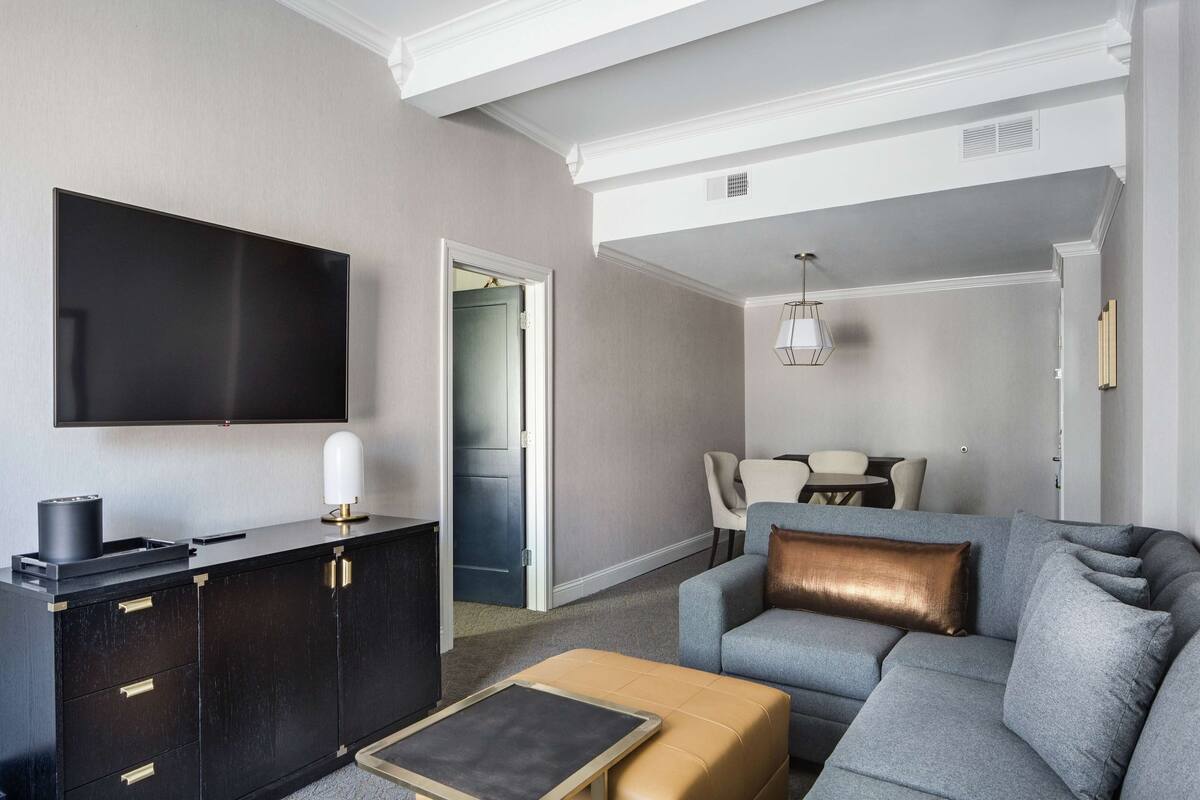 Una sala de estar moderna con un televisor de pantalla plana, un mueble de madera oscura, un reposapiés color beige y un sofá gris con un cojín de color cobre.