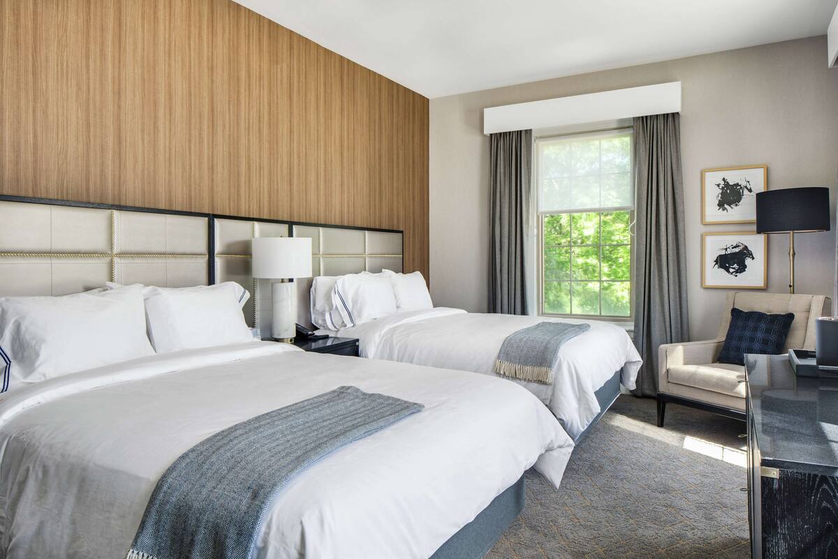 Una habitación de hotel con dos camas, un escritorio, una silla, una lámpara y un ventanal con cortinas.
