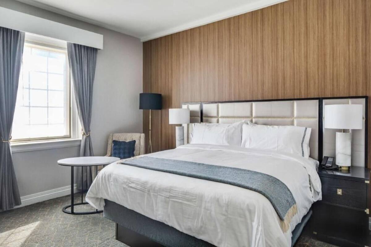 Una habitación de hotel con una cama grande, una mesita redonda pequeña, una lámpara y una mesita de noche.