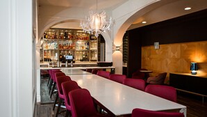 A restaurant with a long white table and red chairs, a bar with shelves of bottles, and a chandelier.