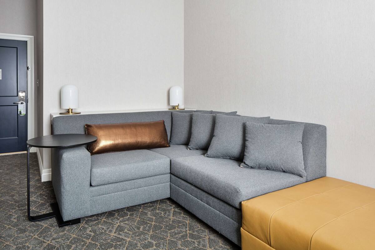 Una habitación de hotel moderna con un sofá seccional gris, una mesita redonda pequeña y un otoman color mostaza.