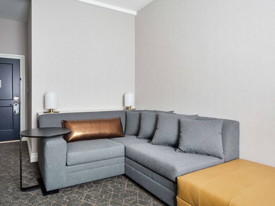 A modern hotel room with a grey sectional sofa, a small round table, and a mustard-colored ottoman.