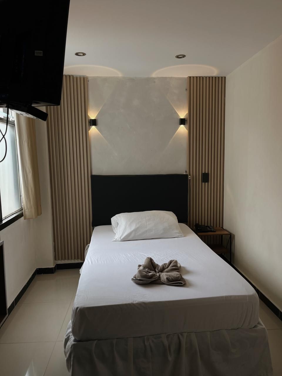 A hotel room with a bed, a bedside table, a TV, and a window with curtains.