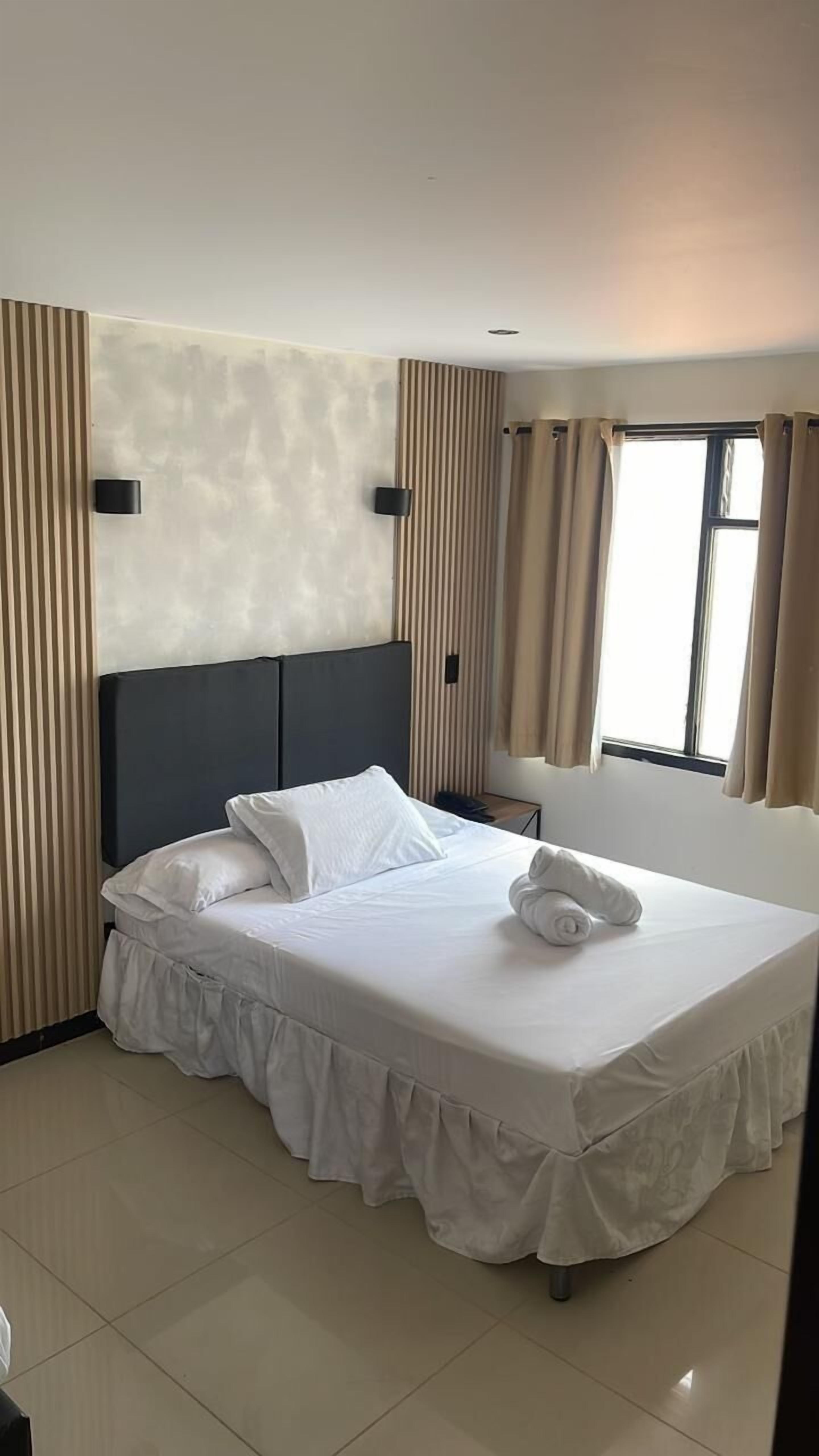 A neatly made bed with white linens and a rolled towel, a bedside table, and a window with curtains.