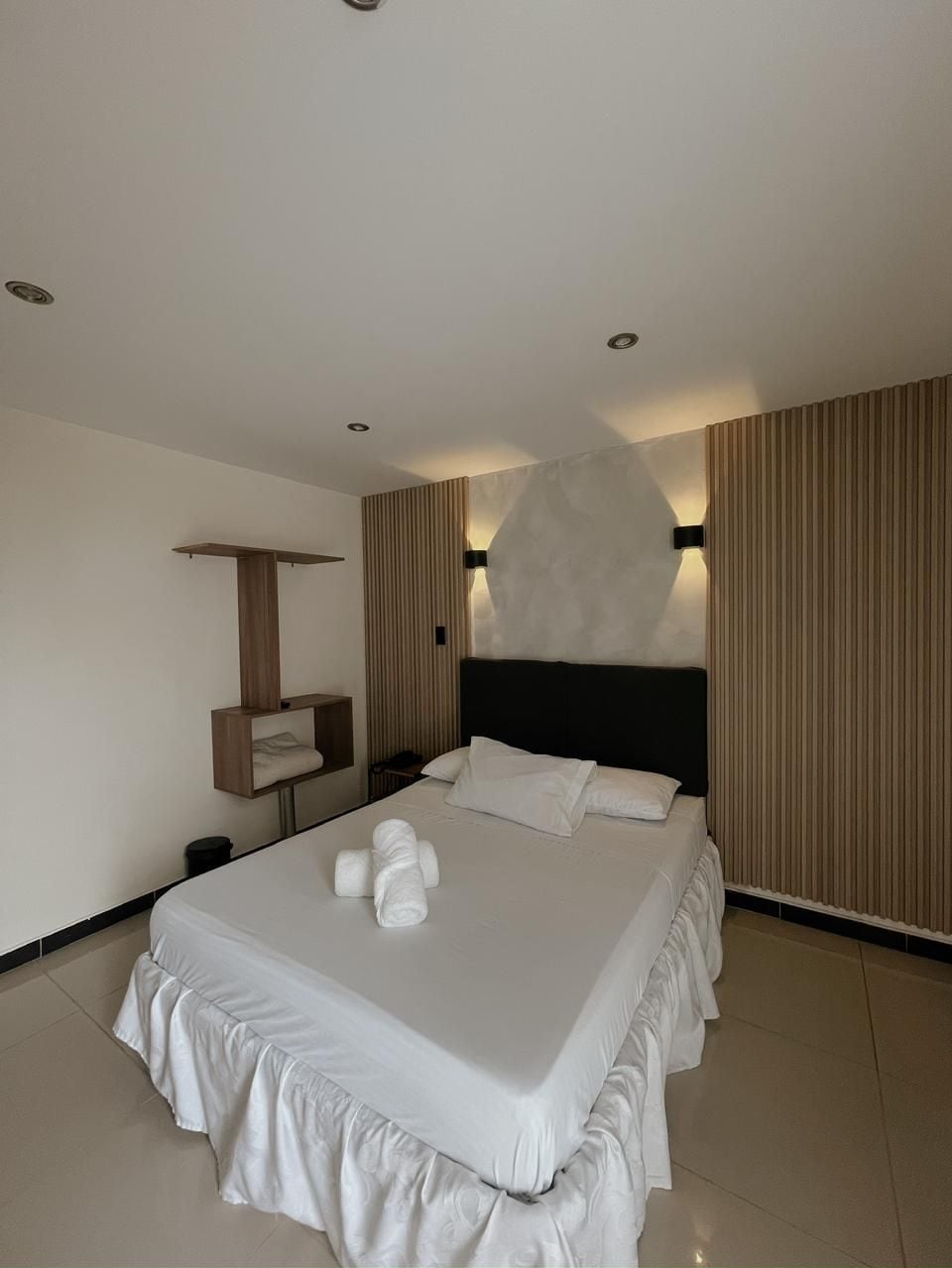A modern hotel room with a large bed, a bedside table, a wall-mounted shelf, and a large window with blinds.