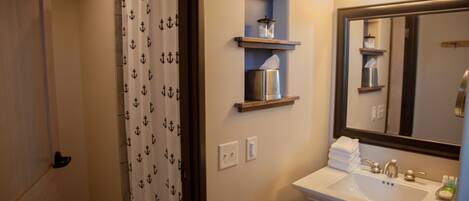 A bathroom with a white sink, a mirror, and a shower curtain with anchor patterns. There are shelves above the sink and a towel rack with folded towels.